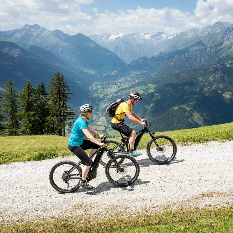 Zwei Personen auf E-Mountainbikes auf einem breiten Weg am Klaunzerberg. Im Hintergrund erstreckt sich das Tal und die umliegende Berglandschaft.