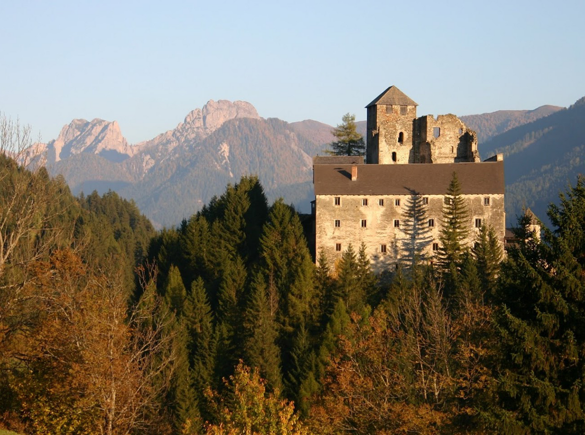 Herbstansicht der mittelalterlichen Burg Heinfels in Sillian, die mächtig vor einer imposanten Bergkulisse thront und unterhalb von herbstlich gefärbtem Wald umgeben ist.