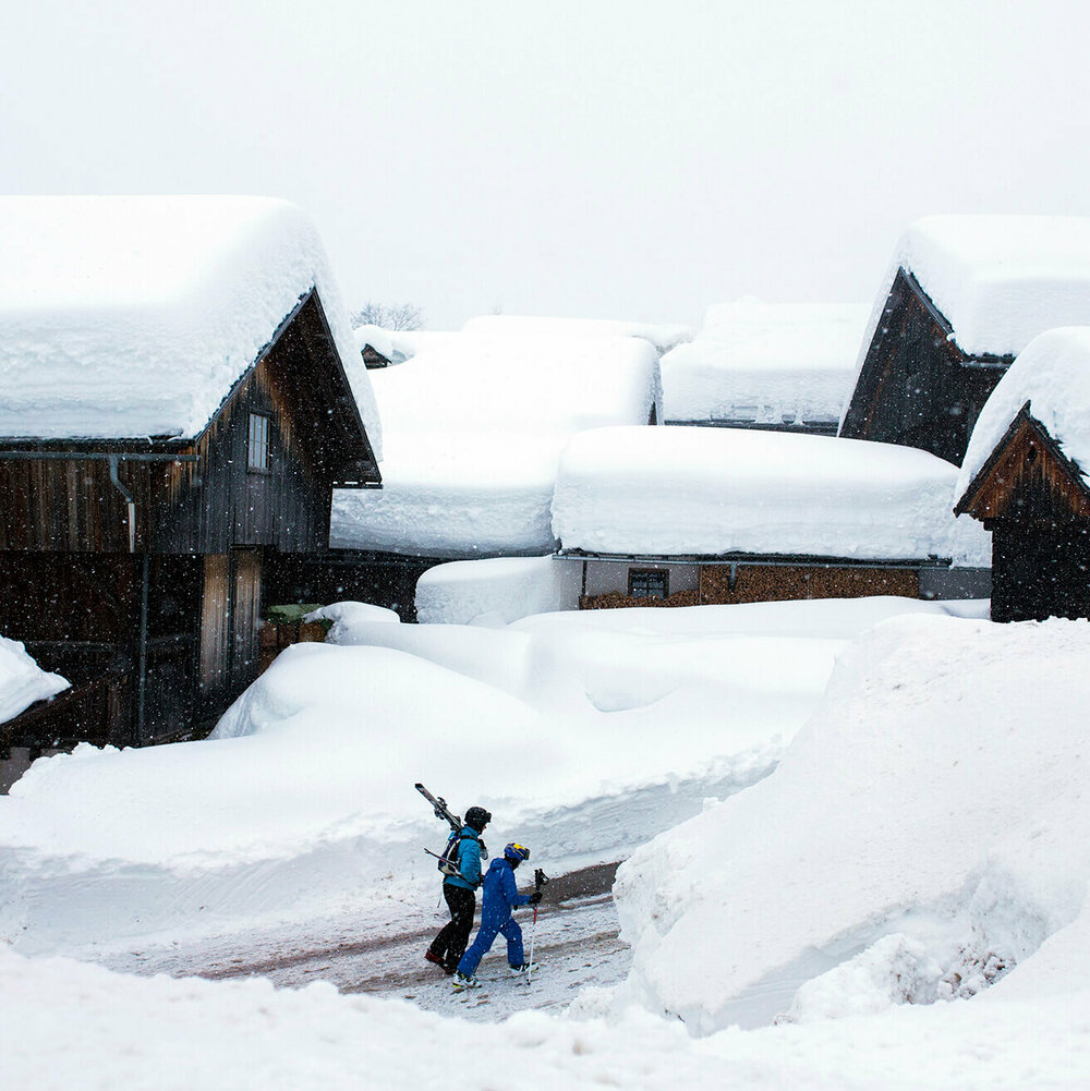 Zwei Skifahrer spazieren in voller Ausrüstung bei Schneefall durch die engen Straßen Obertilliachs, die von hohen Schneewänden umgeben sind.