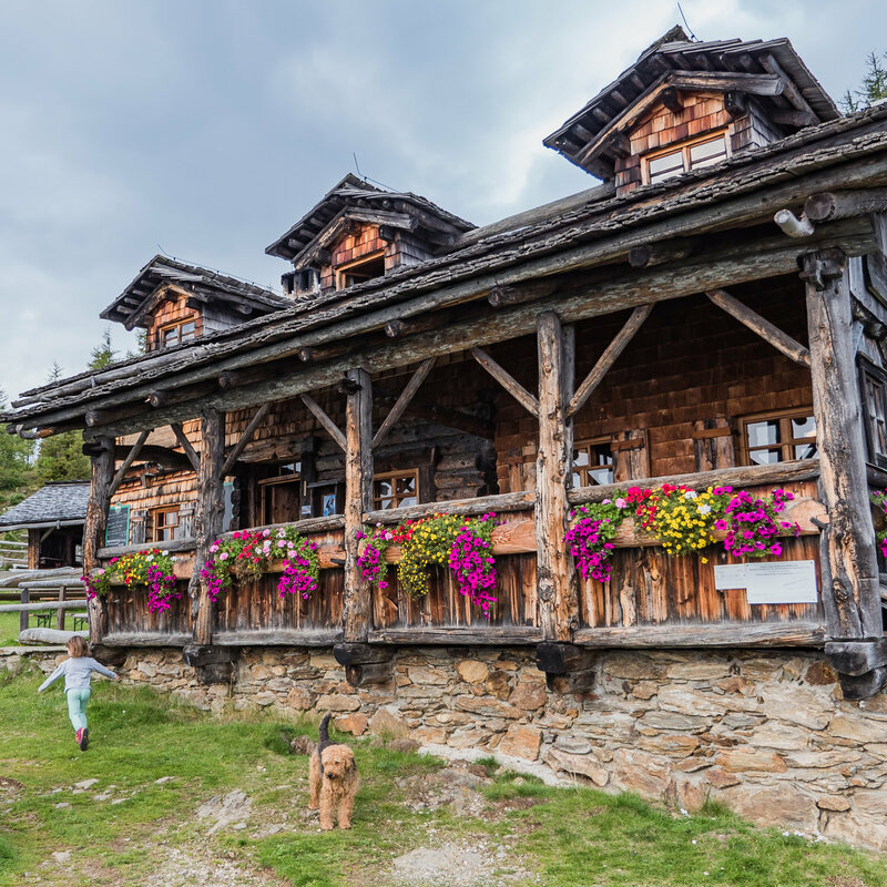 Das urige Anna Schutzhaus aus Holz mit Blumendeko und spielenden Kindern und einem Hund davor.