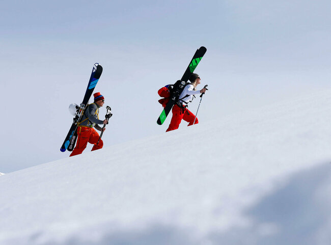 Ein Pärchen steigt mit Ski am Rücken einen schneebedeckten Berg hinauf. Beide tragen Mützen und rote Hosen. Die Ski sind schwarz-grün und schwarz-blau und sind an einem Rucksack befestigt.