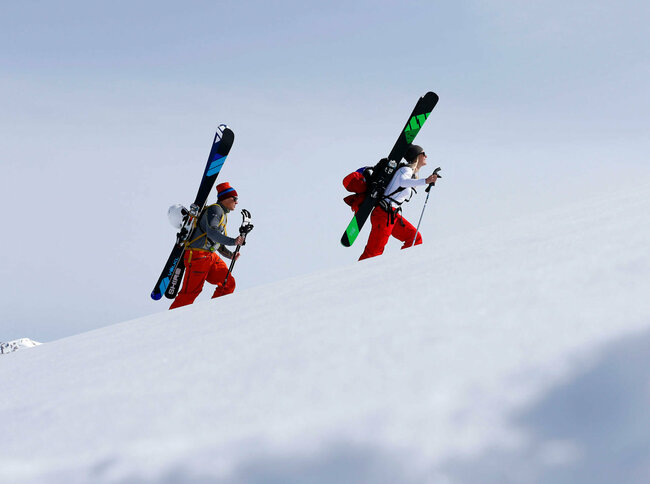 Ein Pärchen steigt mit Ski am Rücken einen schneebedeckten Berg hinauf. Beide tragen Mützen und rote Hosen. Die Ski sind schwarz-grün und schwarz-blau und sind an einem Rucksack befestigt.