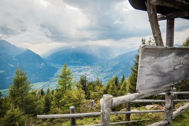 Weite Panoramaaufnahme aus der Vogelperspektive auf ein grünes Tal mit einer Stadt (Lienz) unter einem dramatisch bewölkten Himmel. Im rechten Vordergrund ist eine hölzerne Informationstafel mit Gravuren der Berggipfel zu sehen, die an einem Pfosten montiert ist. Ein rustikaler Holzzaun und Nadelbäume säumen den Bildrand, während sich im Hintergrund die sanften und schroffen Gipfel der Alpen im Dunst erstrecken.