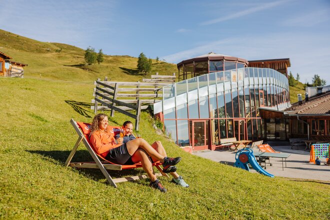 Zwei Frauen liegen in Liegestühlen auf einer grünen Wiese. Dahinter steht ein Gebäude mit Spielplatz.