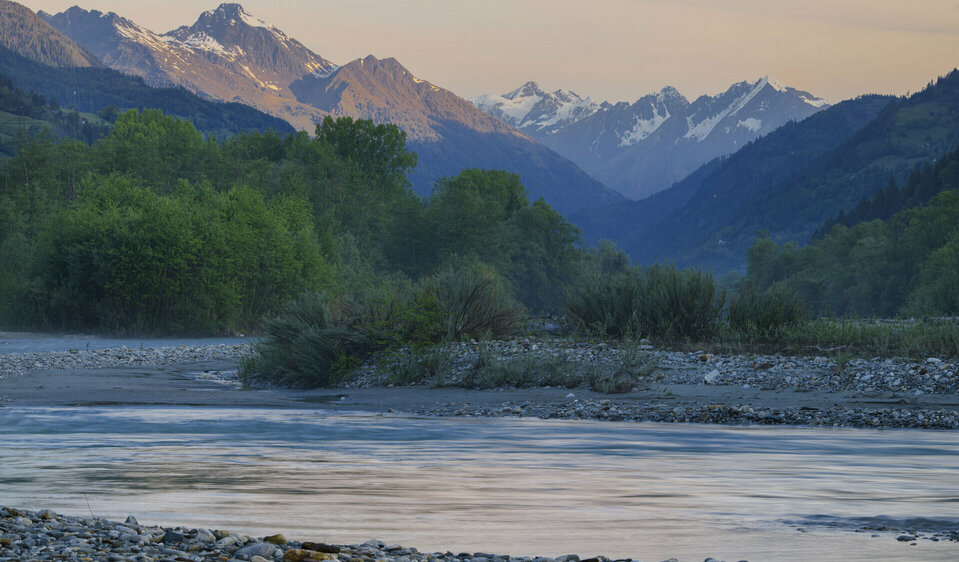 Die Isel macht einen Schwung durch die Landschaft. Auf der Wasseroberfläche spiegelt sich der Himmel wieder. Im Hintergrund sind Berge zu sehen. 