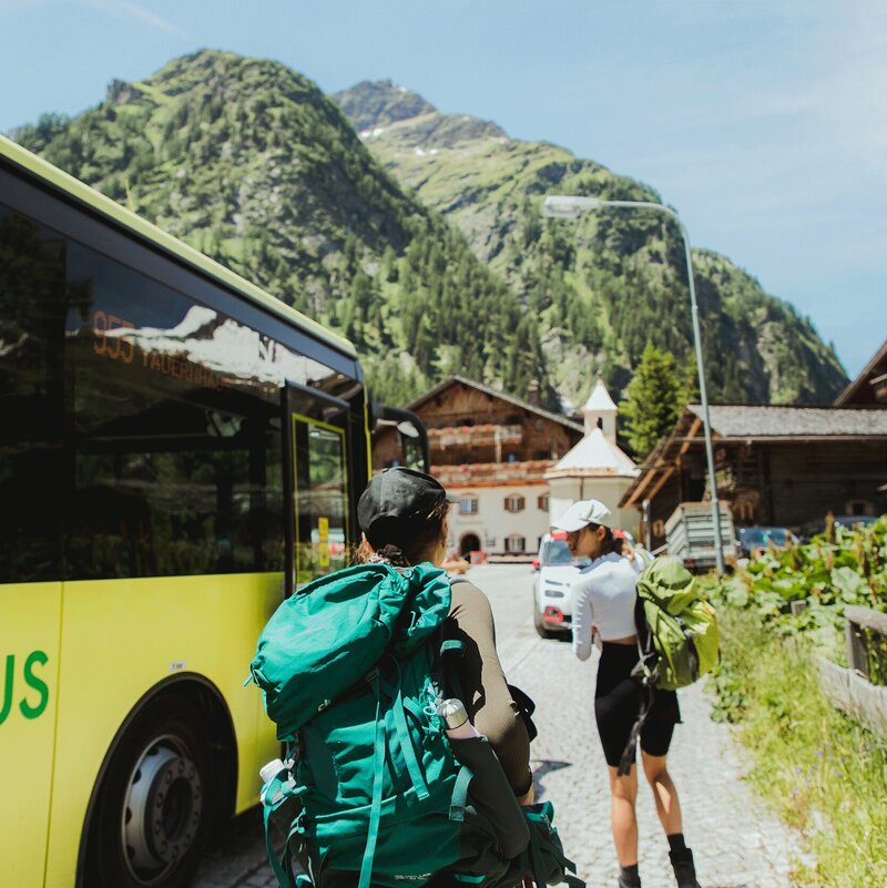 Zwei Wanderer steigen aus dem gelben Linienbus der ÖBB aus, im Hintergrund steht der traditionsreiche Gasthof „Matreier Tauernhaus“ in Matrei in Osttirol.
