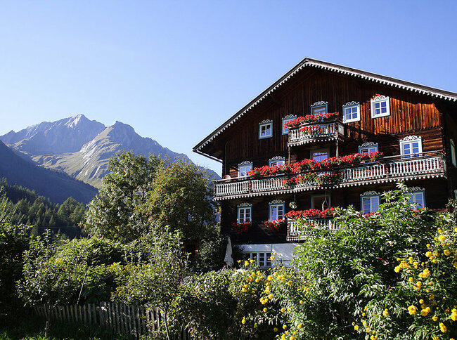 Blick auf ein altes Bauernhaus in Kals mit roten Blumen auf den Balkonen und einem Berg im Hintergrund.
