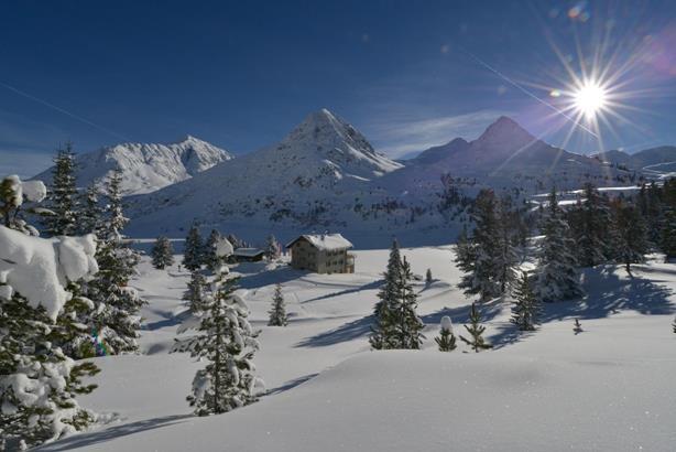 Verschneites Alpengasthaus vor hohen Berggipfeln unter blauem Himmel mit strahlender Sonne.