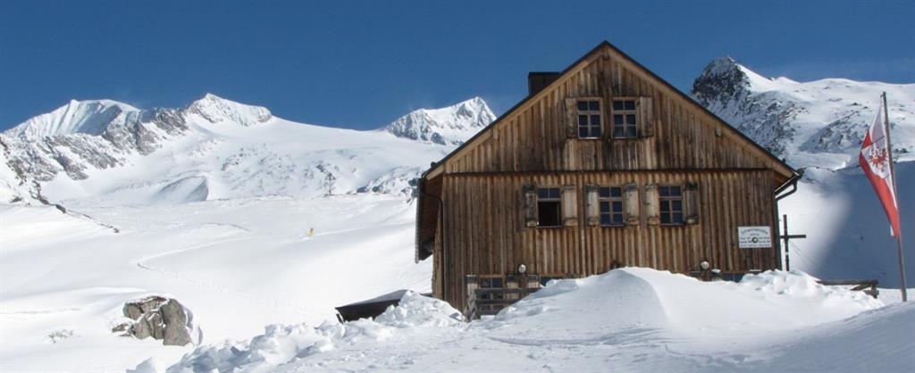 Das Bild zeigt die Johannishütte in einer verschneiten Hochgebirgslandschaft. Die Hütte mit ihrer dunklen Holzfassade steht umgeben von hohen Schneemassen vor einer Kulisse aus schneebedeckten Bergen. Rechts neben dem Gebäude ist eine Fahne zu sehen, darüber spannt sich ein klarer blauer Himmel. 