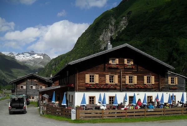 Das Bild zeigt den Alpengasthof Venedigerhaus Innergschlöß in einem alpinen Tal. Vor dem Gebäude befinden sich eine Terrasse mit mehreren blauen Sonnenschirmen, ein Holzzaun und ein Weg. Im Hintergrund sind ein bewaldeter Berghang sowie Berge mit Schneefeldern zu sehen.